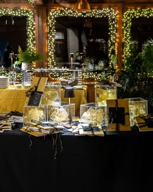 Tavolo decorato con scatole regalo trasparenti illuminate, quaderni dorati con nastri neri e luci calde di Natale in una sala accogliente con cornici di luci sulle finestre e tovaglie giallo ocra.