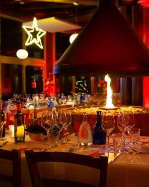 Tavolo apparecchiato con eleganza attorno a un camino centrale acceso, circondato da bottiglie di vino, bicchieri di cristallo e decorazioni natalizie calde con luci rosse soffuse e una stella luminosa sospesa.