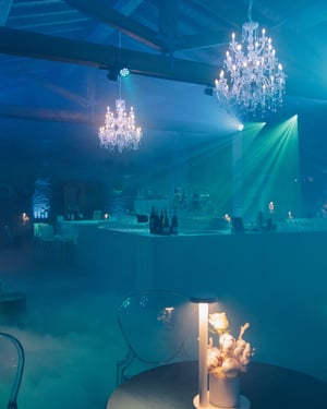 Ambiente elegante e avvolgente con luci blu, lampadari di cristallo, nebbia artificiale e tavoli arredati con fiori bianchi e lampade minimal. Il bar centrale è illuminato per un’atmosfera da festa sofisticata.